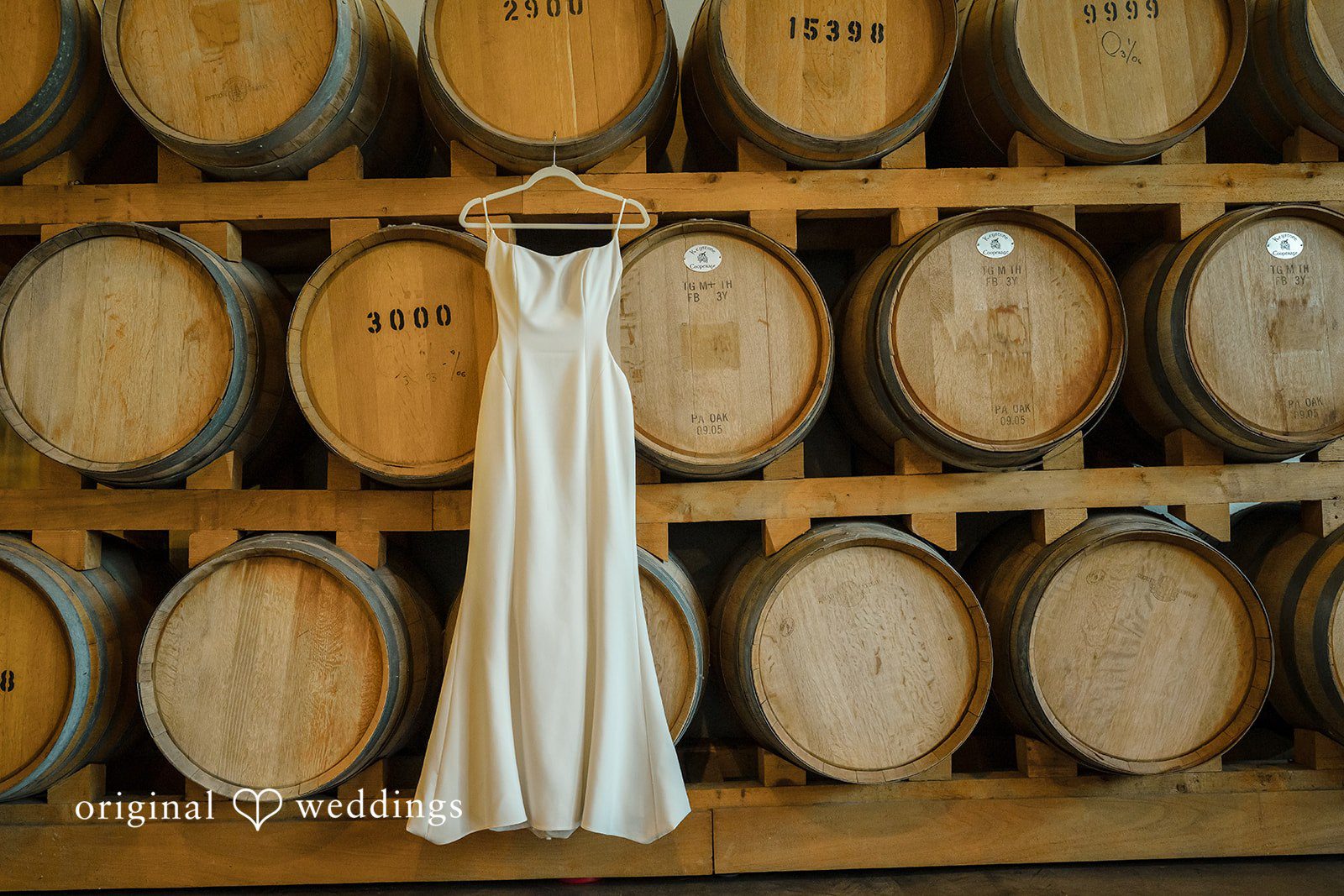 A white wedding dress hanging on a wooden rack in front of stacked wine barrels.