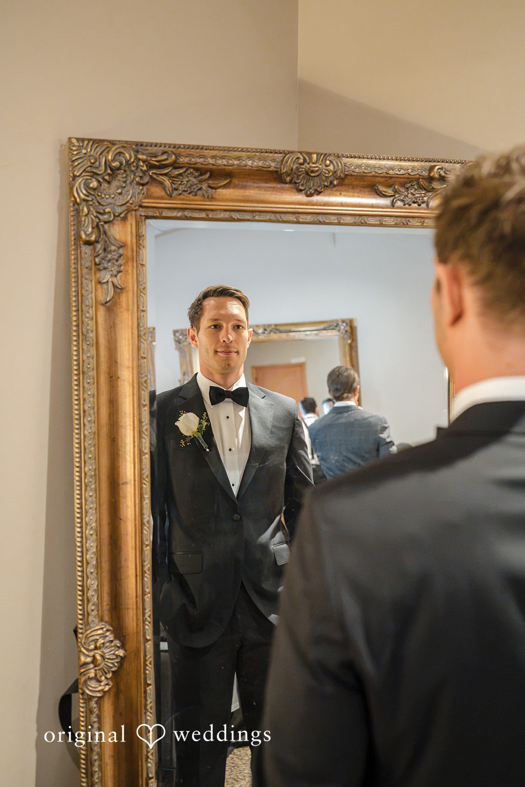 A groom looking at his reflection in a large, gold-framed mirror.