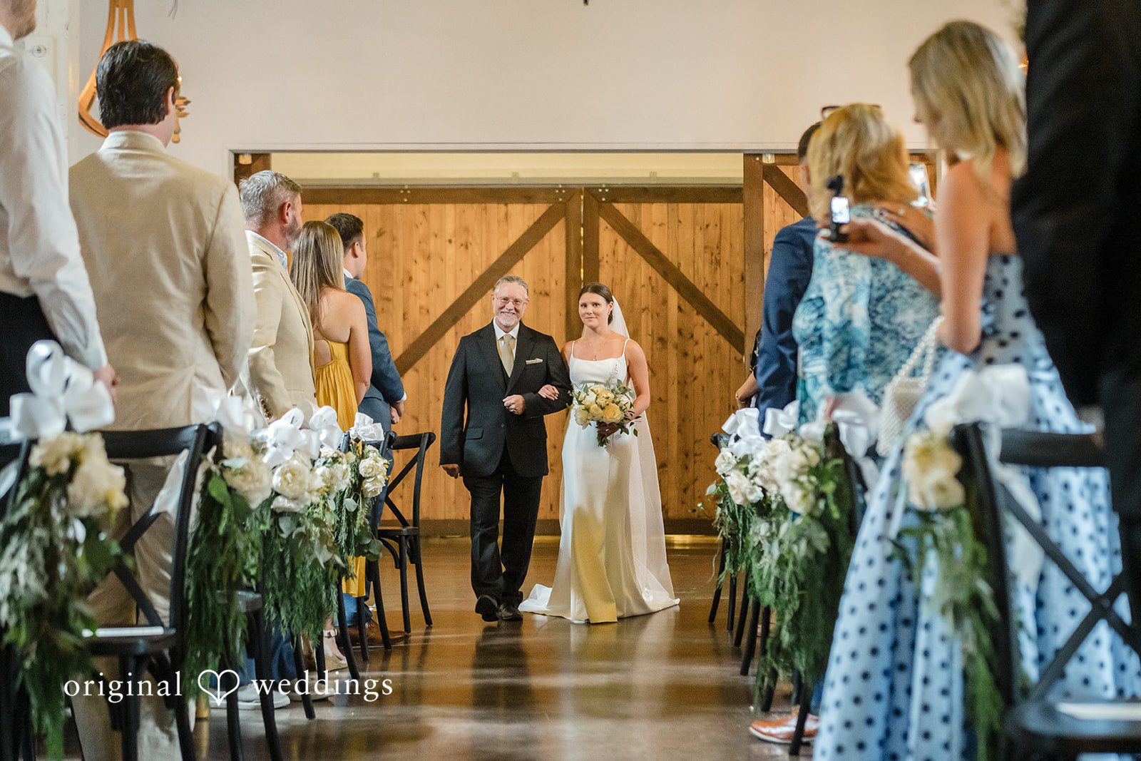 A bride in a white wedding gown walks down the aisle arm-in-arm with an older man in a suit while guests watch from their seats.