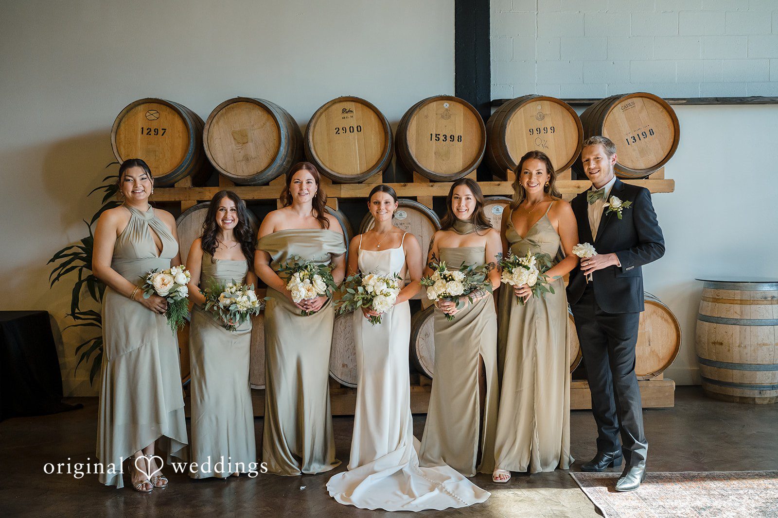 A full wedding party consisting of bridesmaids and man posing together in front of wine barrels.