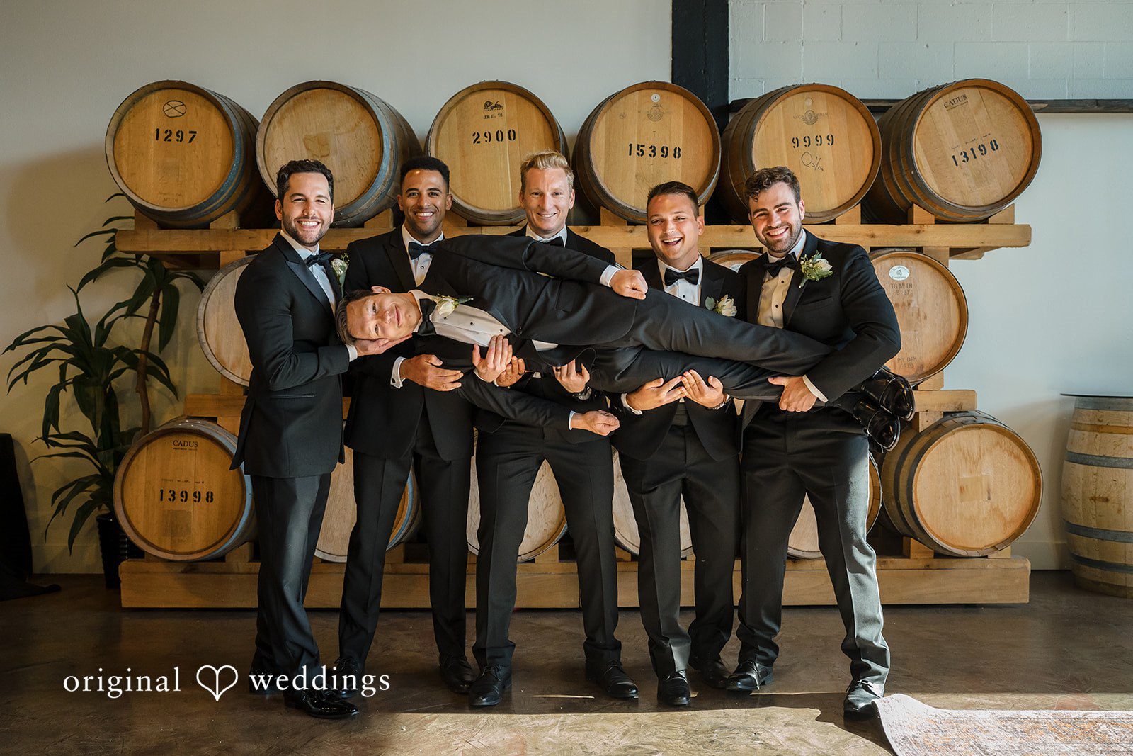 Groomsmen in dark suits standing together and laughing in front of wine barrels.