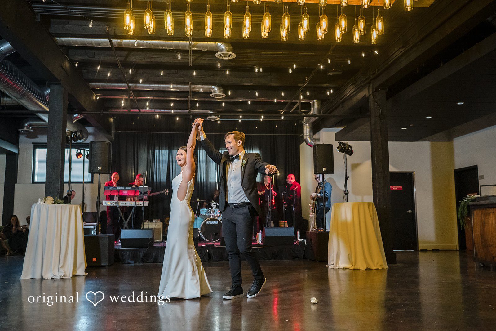 A bride and groom smile joyfully while holding hands high during their dance in front of a live wedding band.