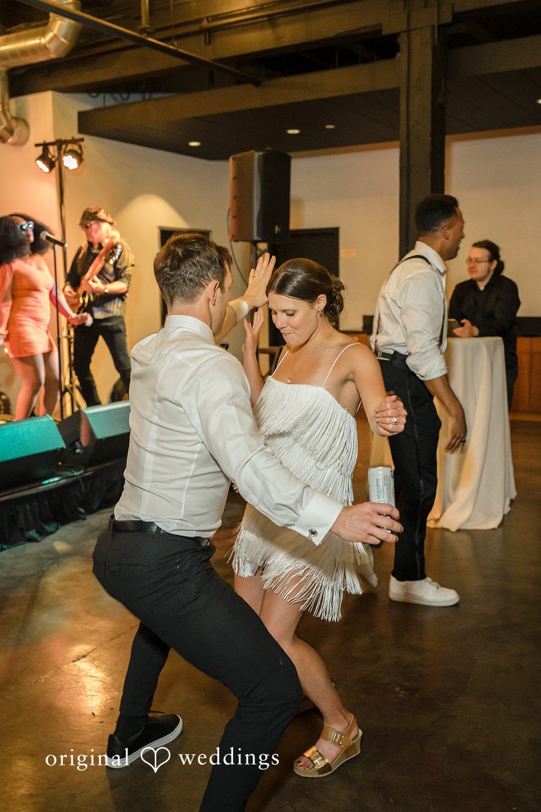 A bride in a short, fringed dress and a man in a white button-down shirt dance energetically together.