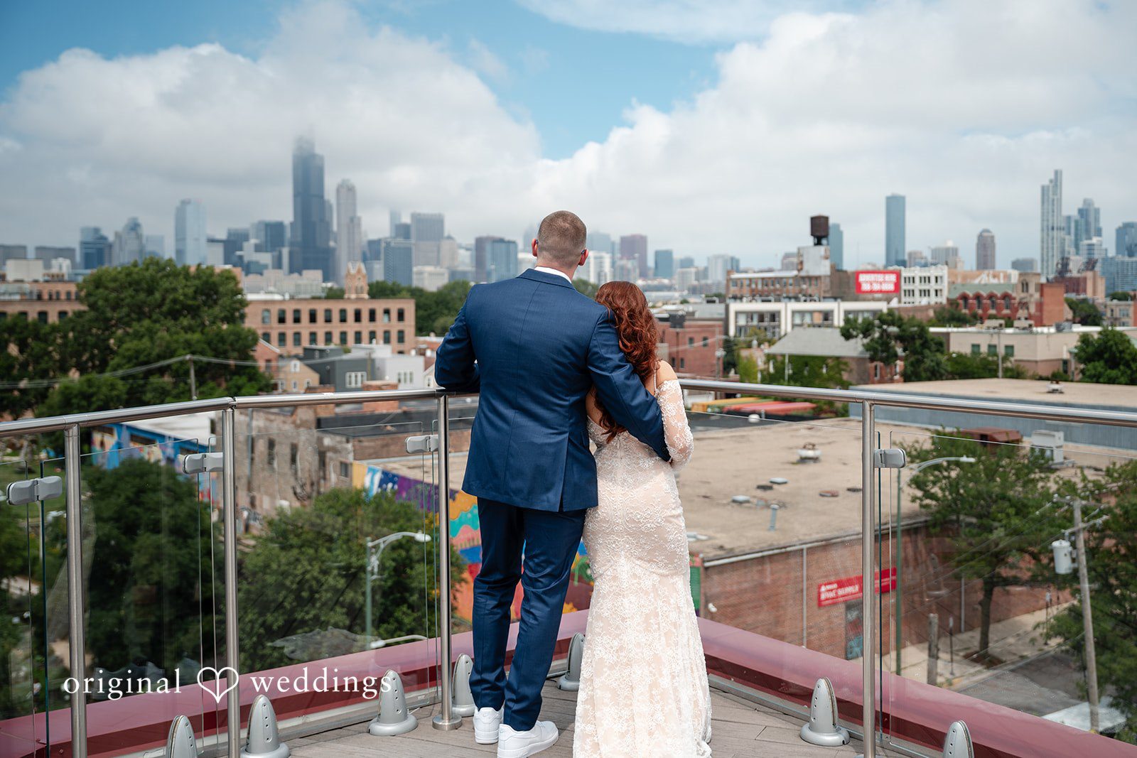 Lucy + Cormac A bride and groom walking hand-in-hand along a rooftop terrace railing.