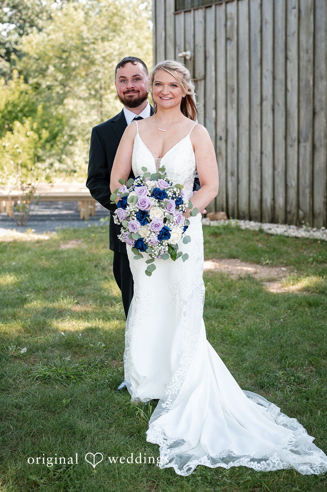 A bride and groom posing while standing on a grassy lawn.