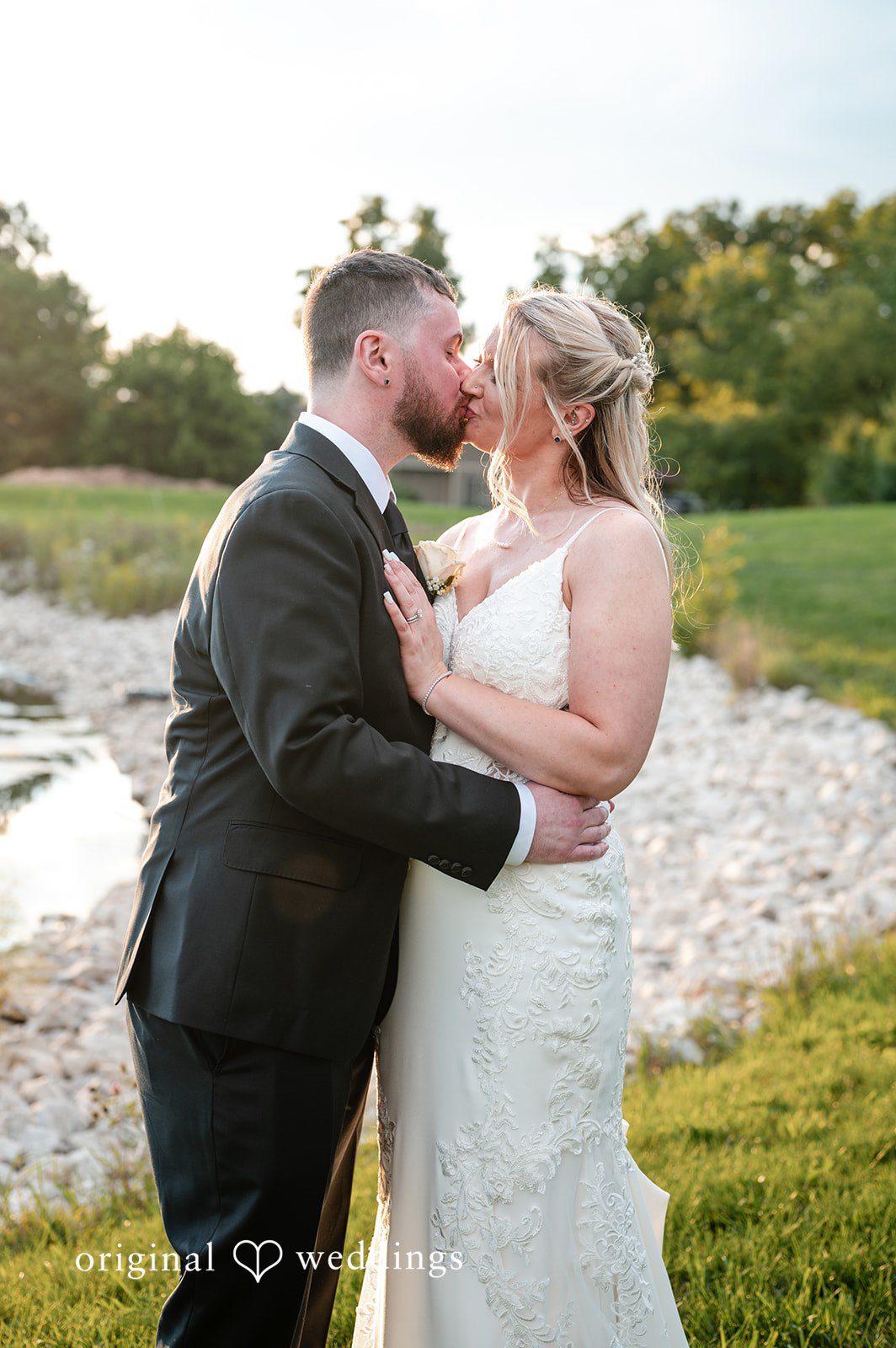 A bride and groom embracing closely near a grassy hillside.