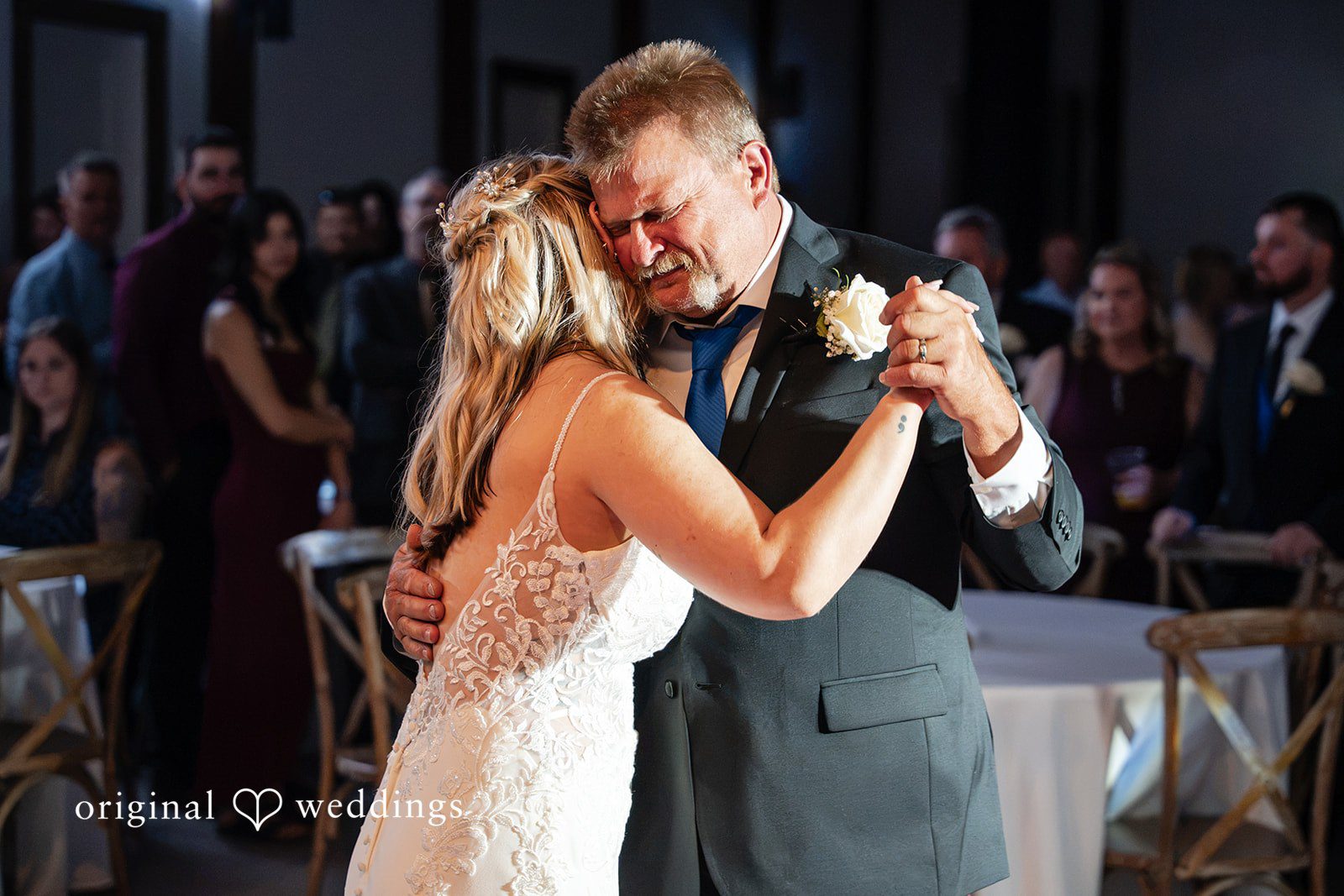 A bride and an older man sharing an emotional dance together while wedding guests watch in the background.