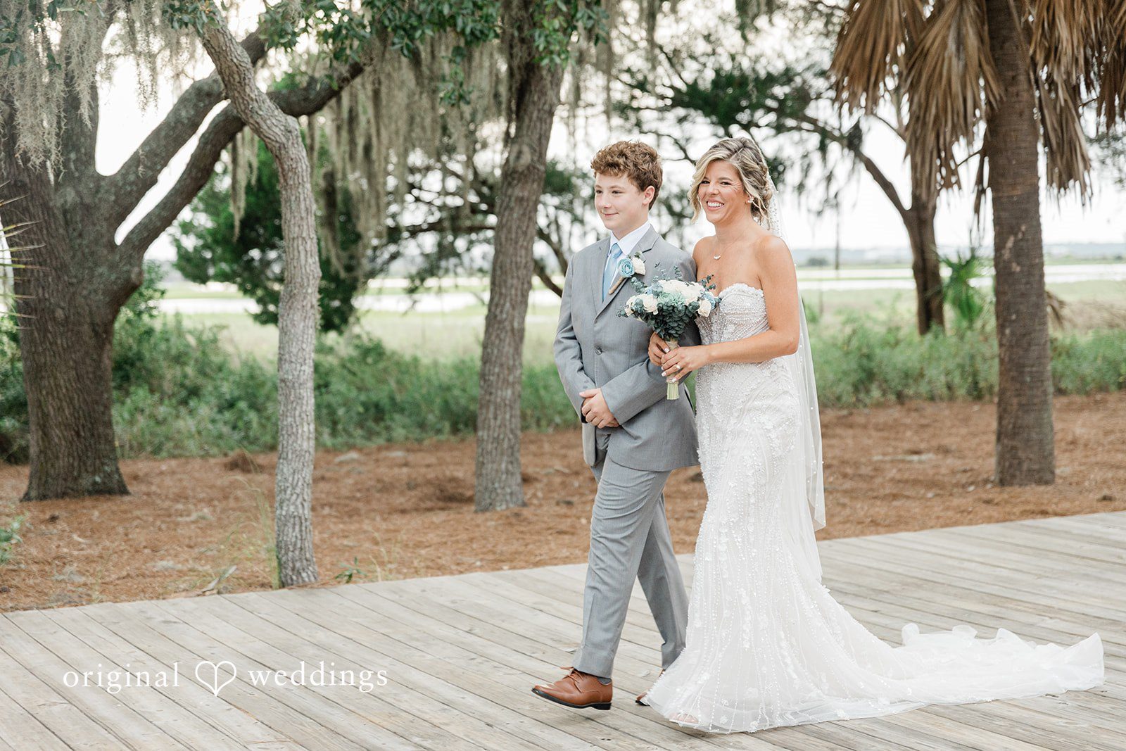 Jennifer + Zac A young boy in a grey suit walking down a path next to his bride in a white gown.