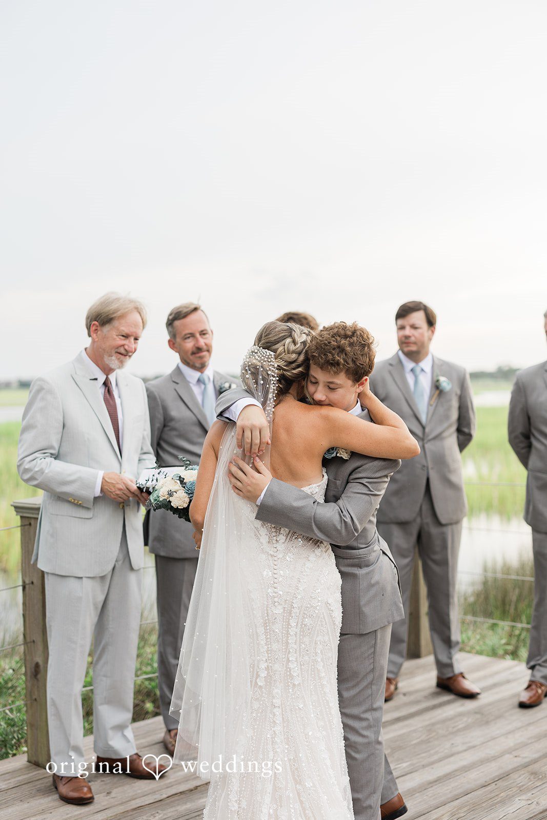 Jennifer + Zac A bride in a lace wedding gown hugs a young boy in a grey suit during an outdoor ceremony.