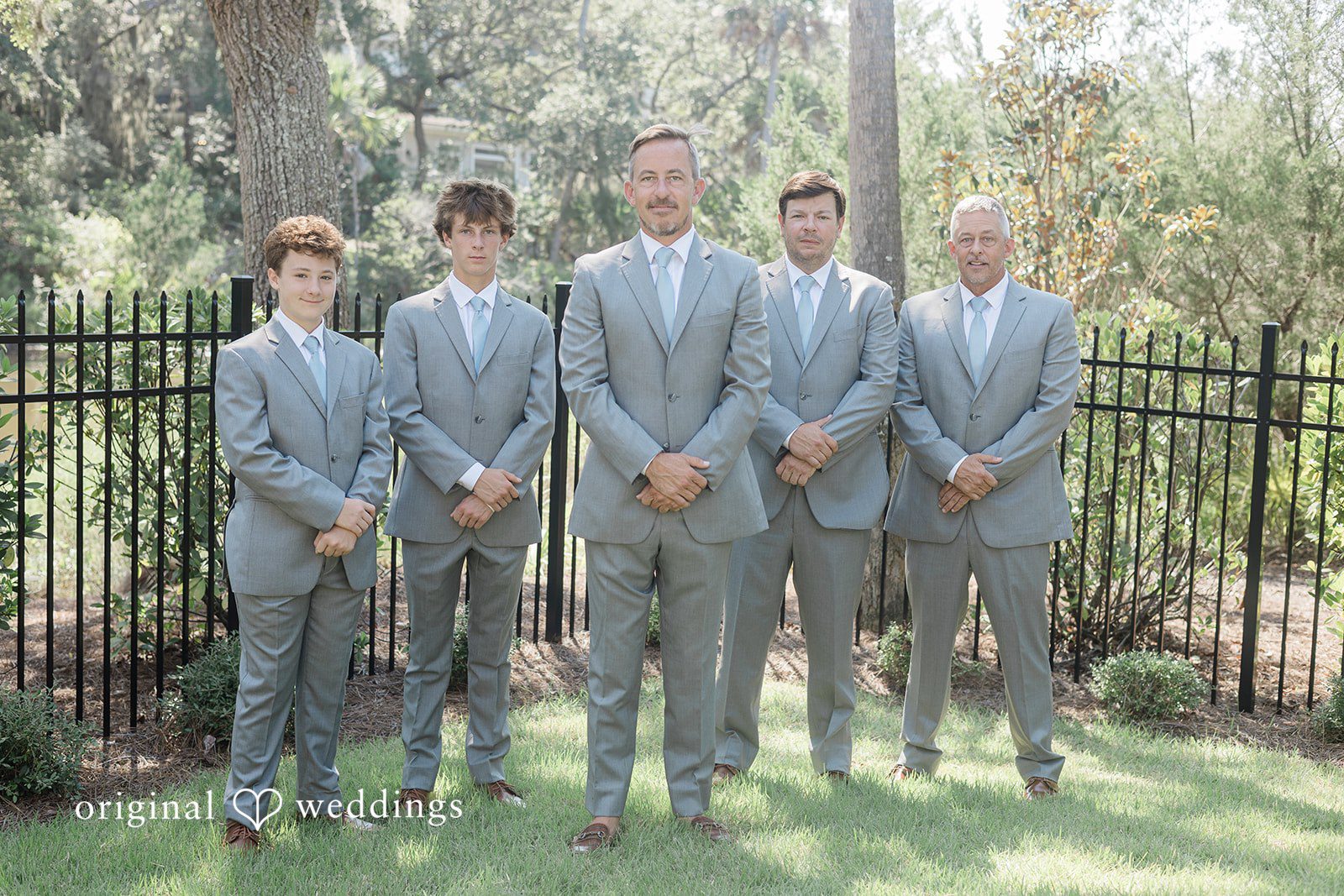 Jennifer + Zac Groomsmen in grey suits posing together against a backdrop of trees and a black fence.