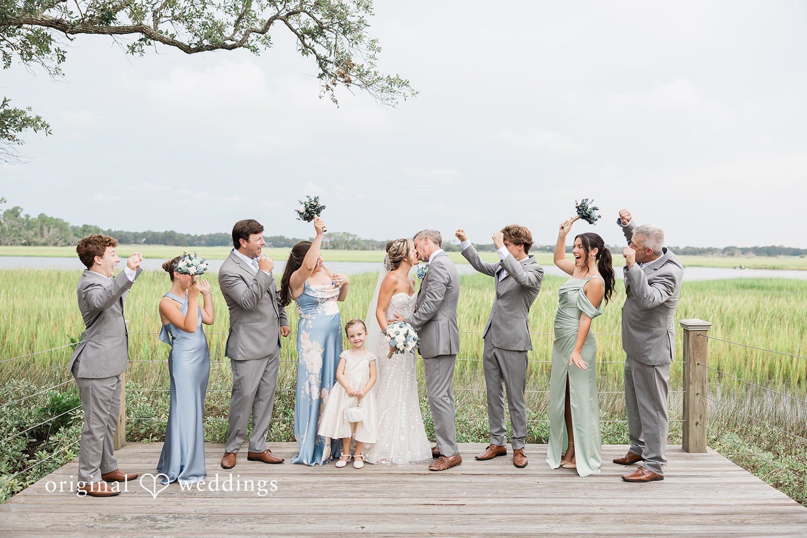 Jennifer + Zac A wedding party raising their hands in celebration during a group photo on a dock.