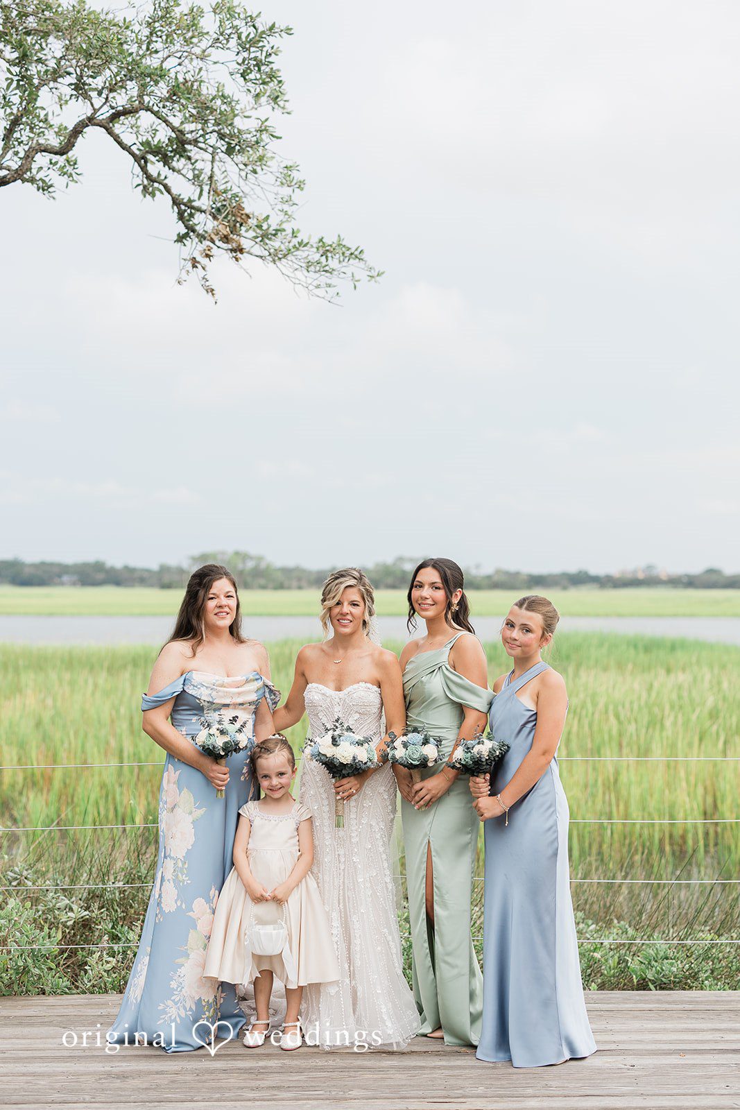 Jennifer + Zac Bride and bridesmaids in shades of blue dresses posing together in an open grassy field.