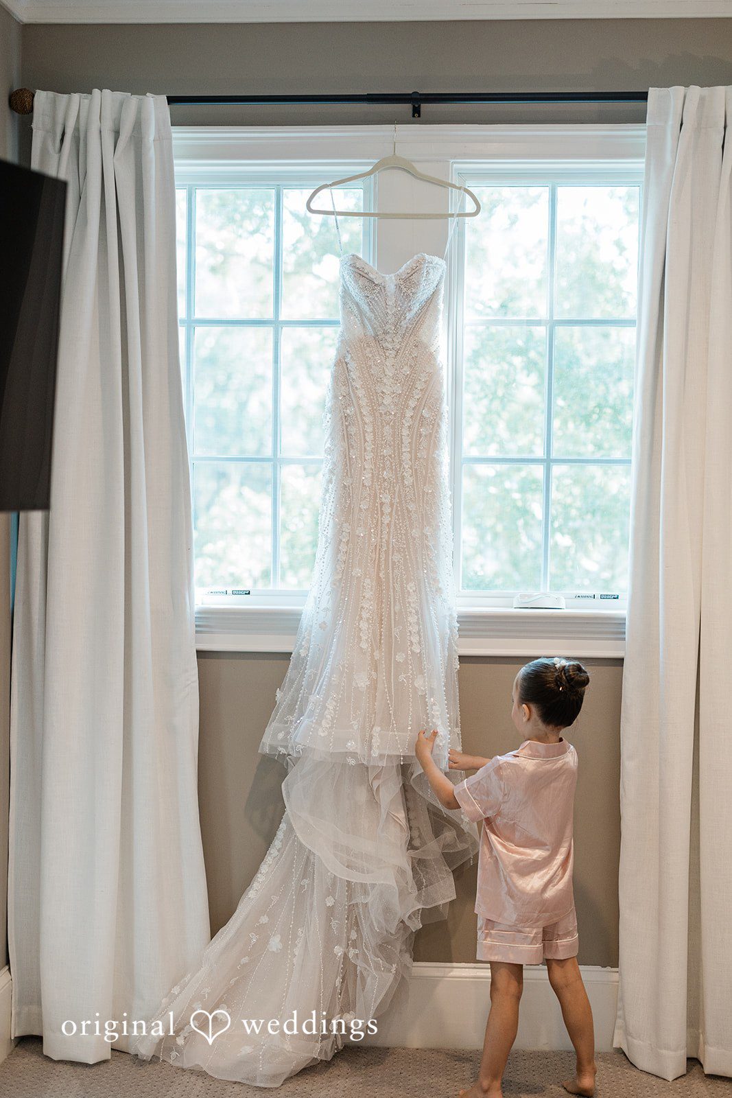 Jennifer + Zac A white wedding dress hanging by a window while a small toddler in pajamas looks up at it.