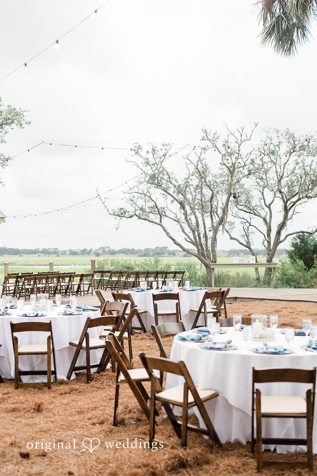 Jennifer + Zac An empty wedding reception setup with rustic wooden chairs and rectangular tables under the trees.