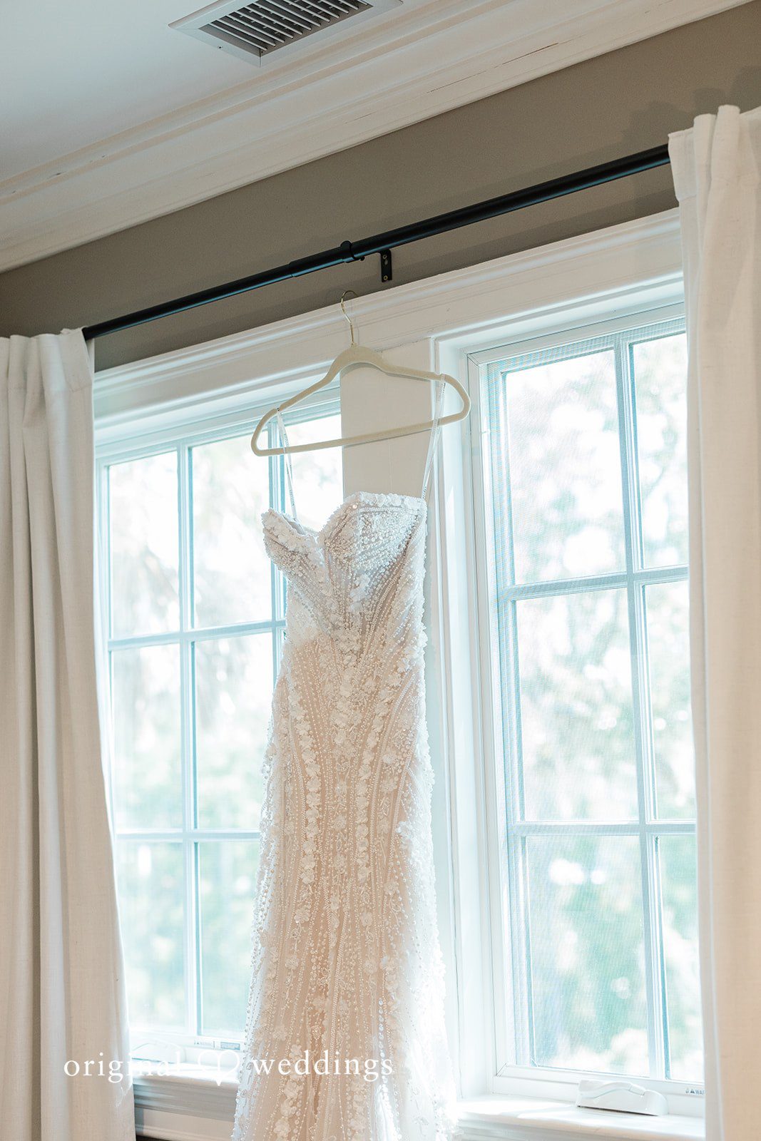 Jennifer + Zac A white lace wedding dress hanging in front of a bright window inside a room.