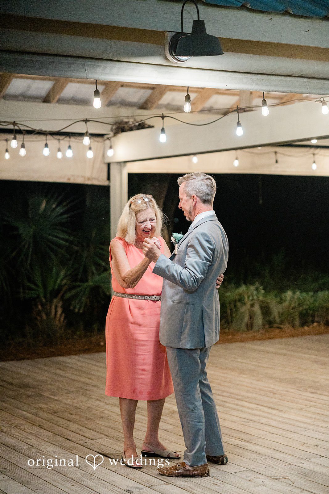 Jennifer + Zac A groom in a suit dancing with a woman in an orange dress on a wooden floor at night.