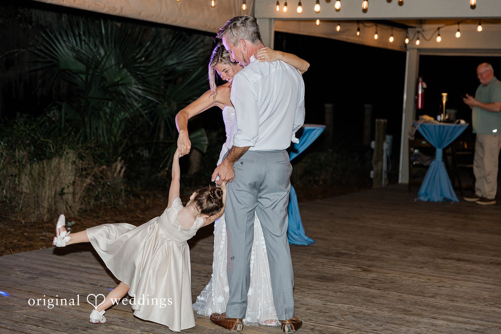 Jennifer + Zac A groom holding a young girl's hand as she dances in a white dress under string lights.
