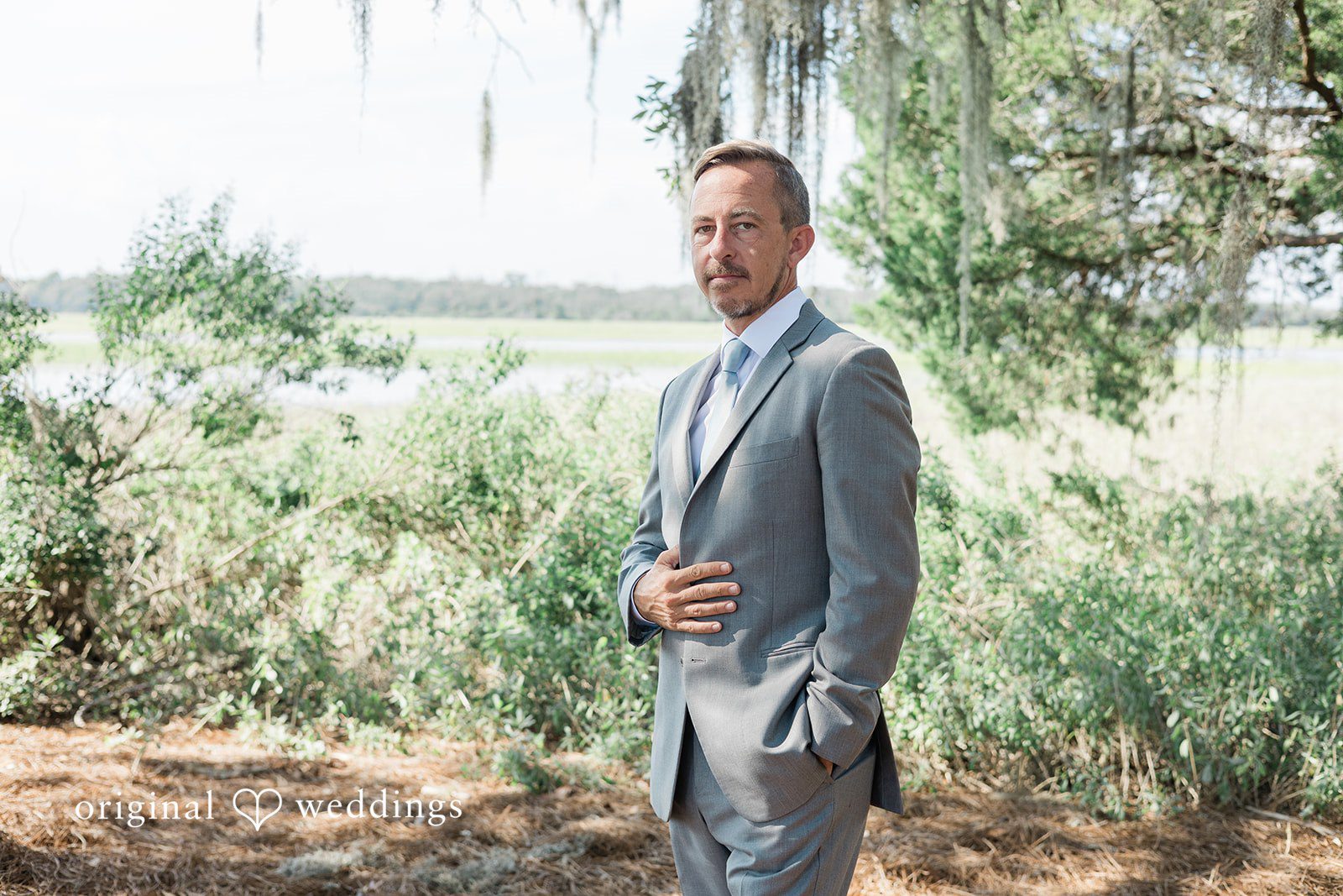 Jennifer + Zac A groom in a light grey suit posing against a background of trees.