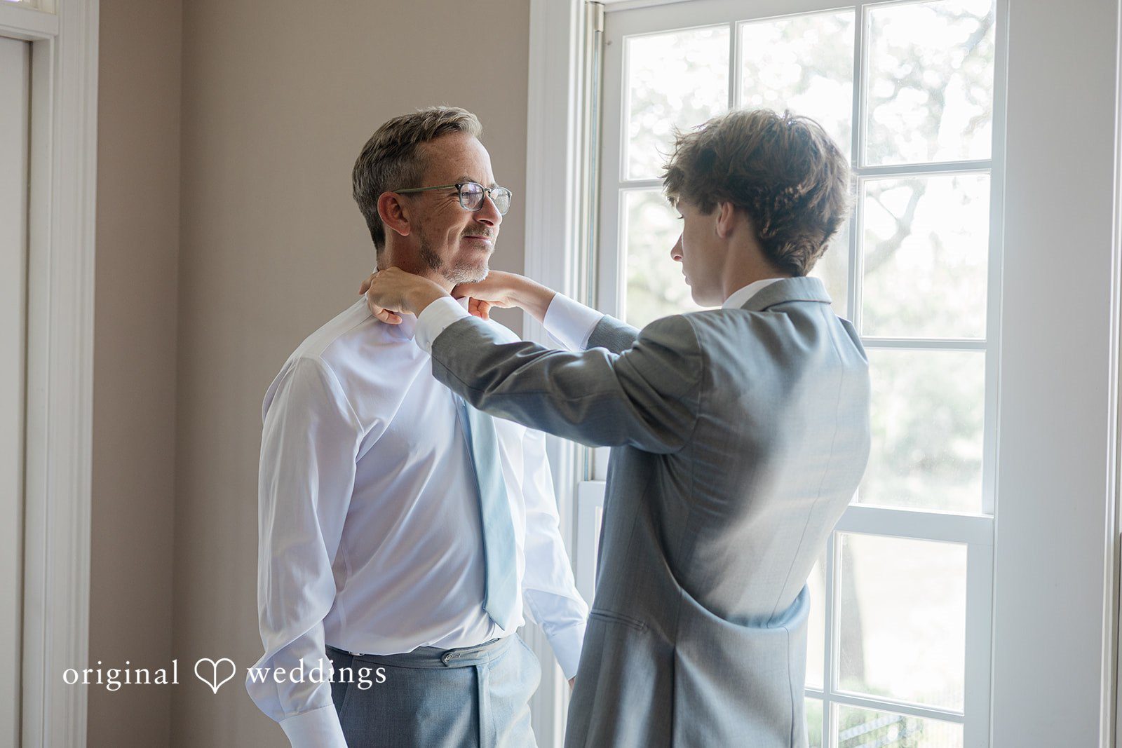 Jennifer + Zac A groom getting his tie adjusted by another man in front of a bright window.