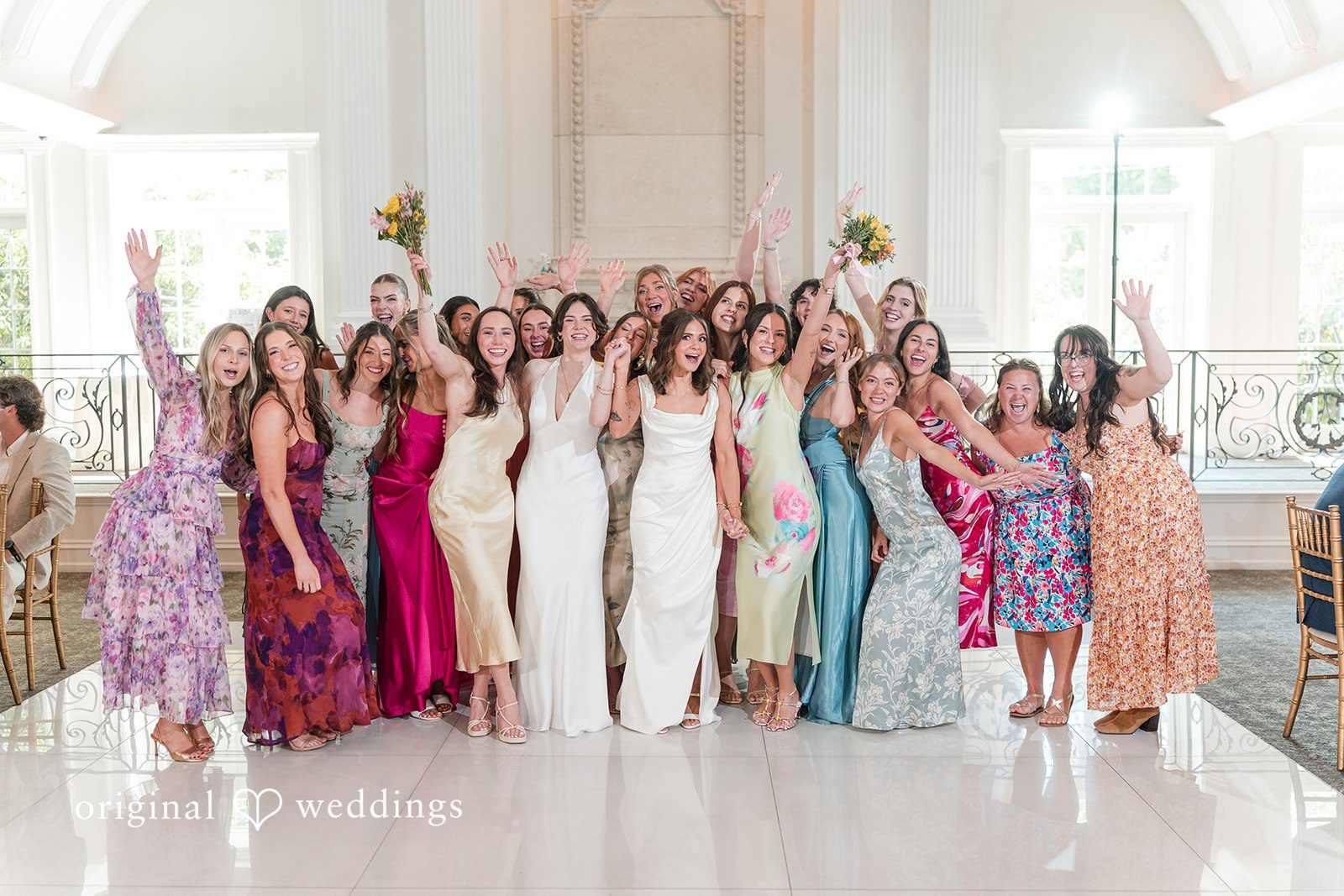 A large group of bridesmaids in vibrant, multi-colored dresses cheering with their hands up.