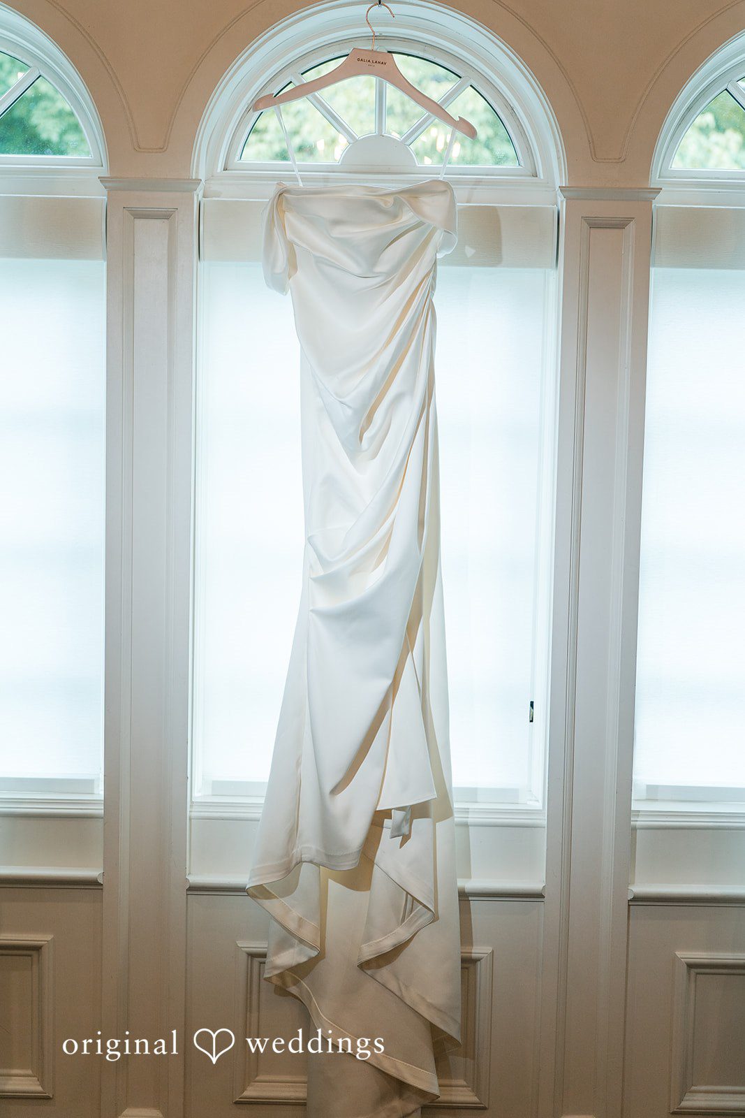 A white wedding gown hanging in front of a bright window.