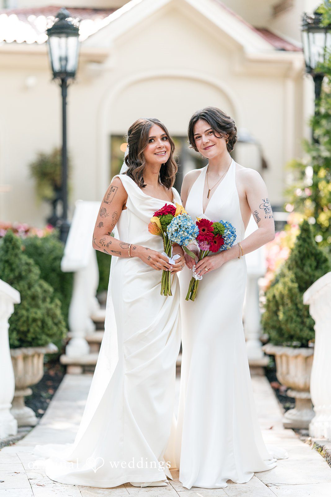 Couple in white dresses smiling and holding flower bouquets together.