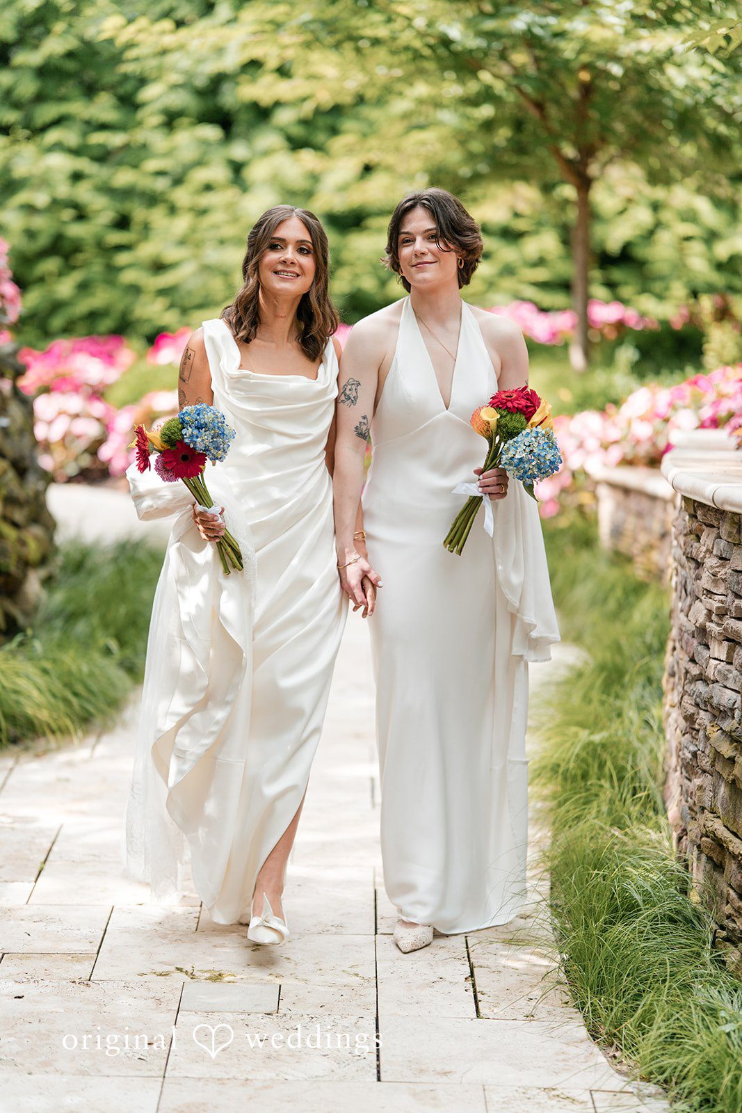 Couple in white gowns holding bouquets and walking together down a pathway.
