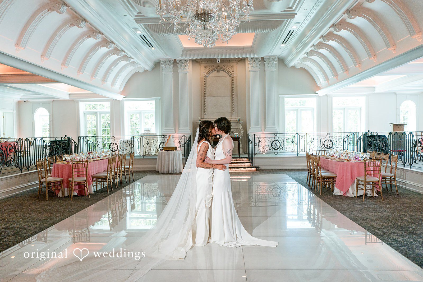 Couple Two brides in white gowns embrace on a polished dance floor in a grand ballroom with a vaulted ceiling and tables set with pink linens.
