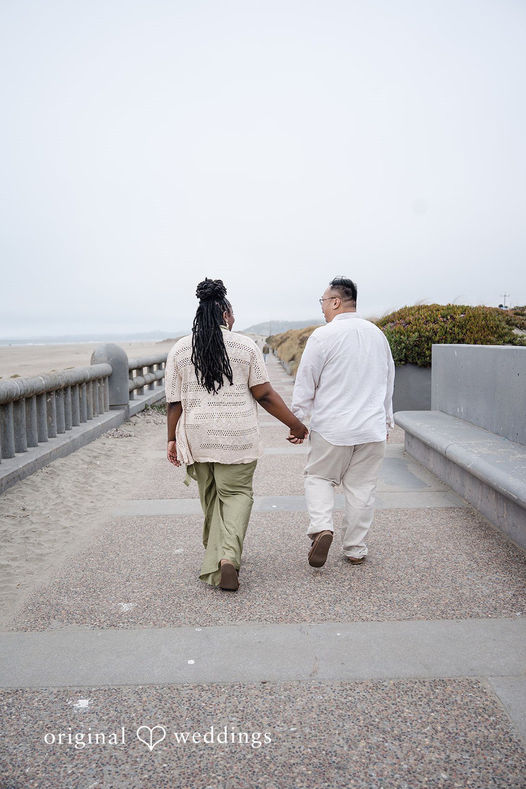 Ashley + Willard Couple holding hands walk away on a paved walkway leading toward a beach.