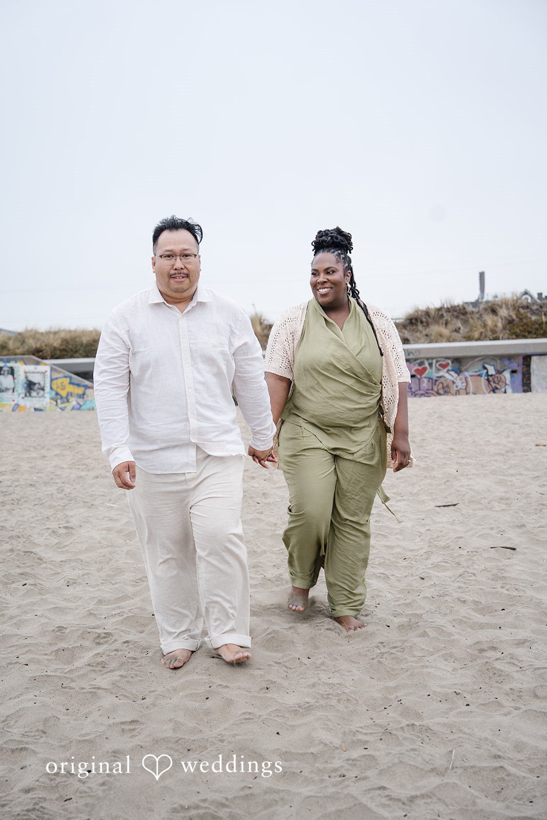 Ashley + Willard Couple walk side-by-side across a sandy beach expanse.