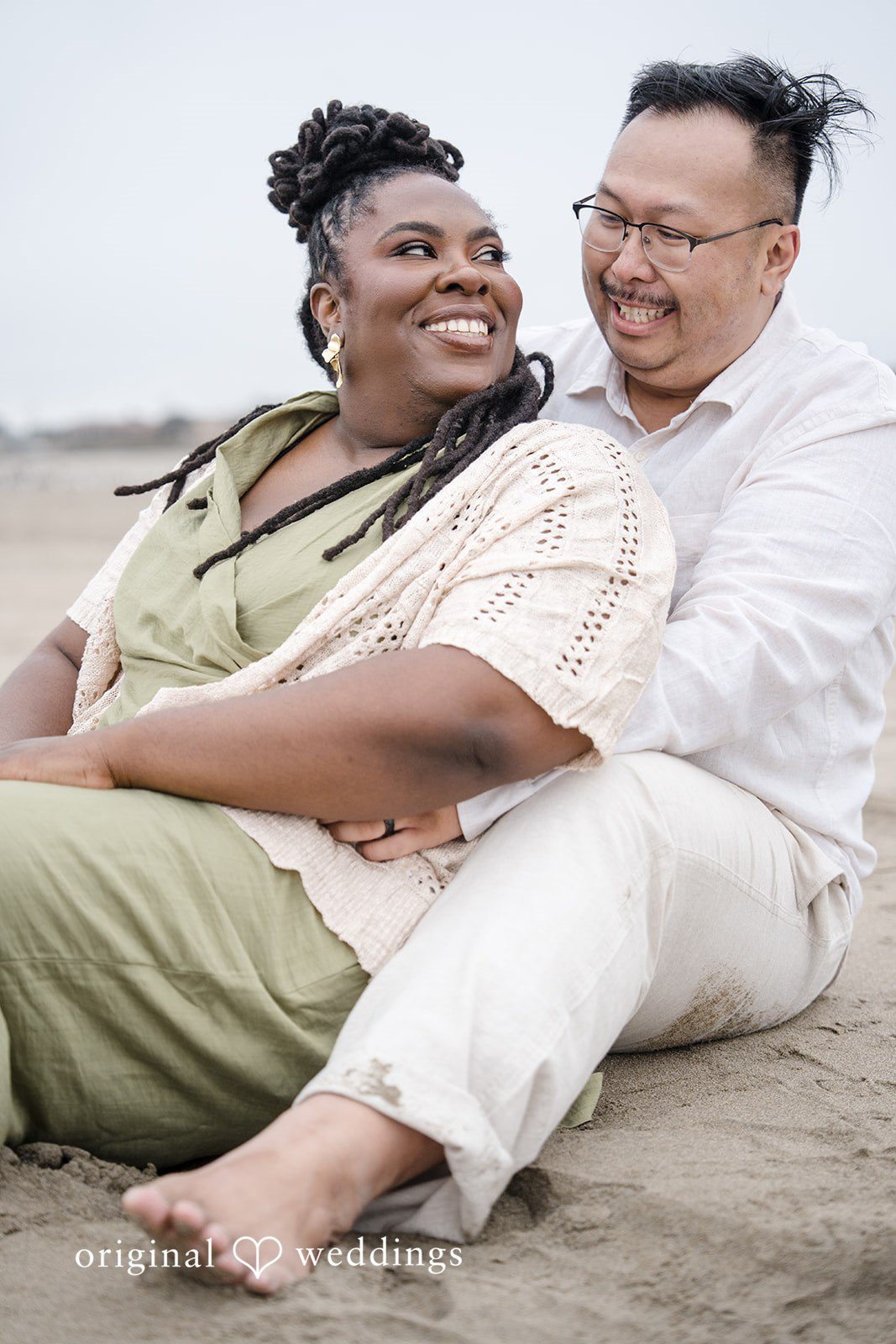 Ashley + Willard One person leans back and smiles happily while sitting against another person on the sand.