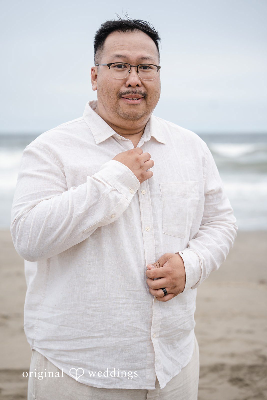 Ashley + Willard A man wearing a white collared shirt adjustments his collar while smiling on a beach.