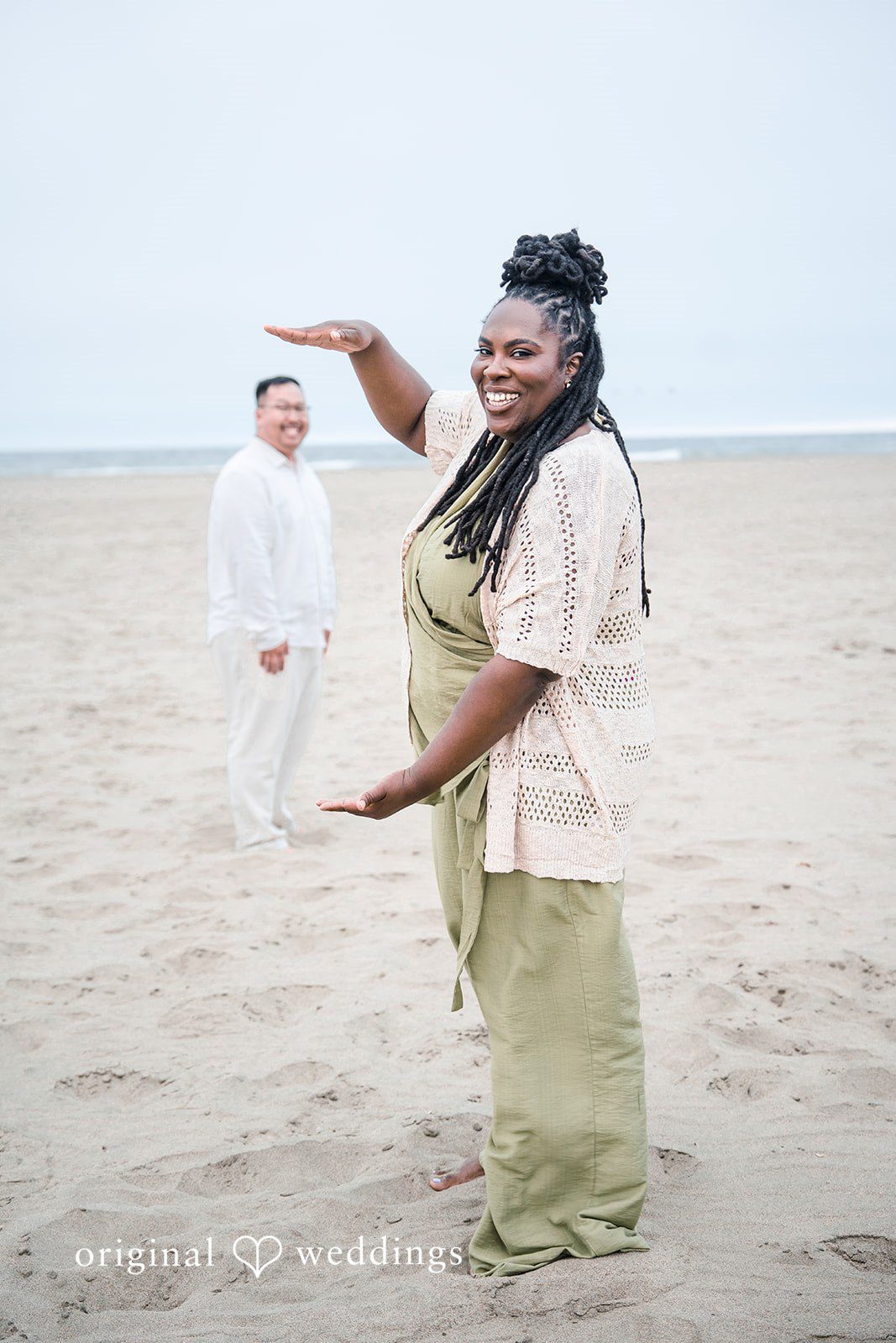 Ashley + Willard One person stands on the beach gesturing toward another individual dressed in white further away.