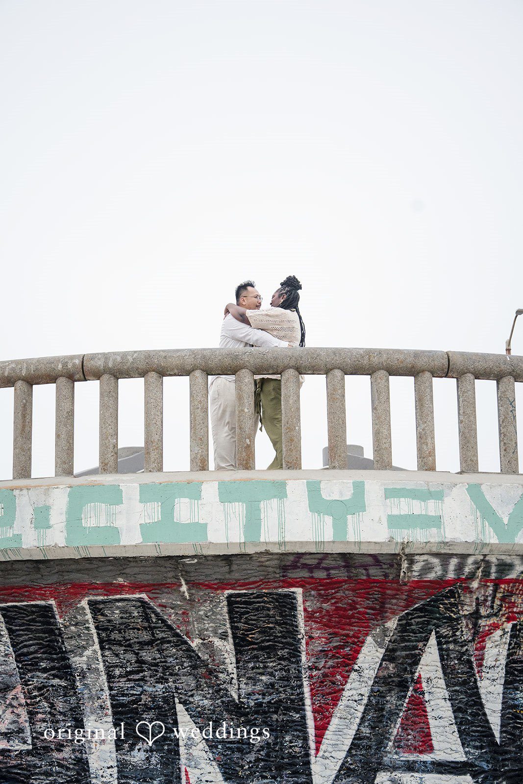 Ashley + Willard Couple stand together on top of an elevated concrete platform looking out.