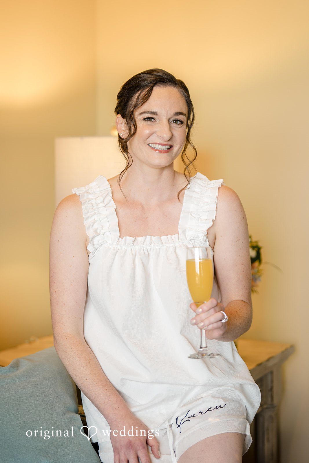 Daniel + Karen A bride in a white robe sitting on a bed smiling while holding a glass of champagne.