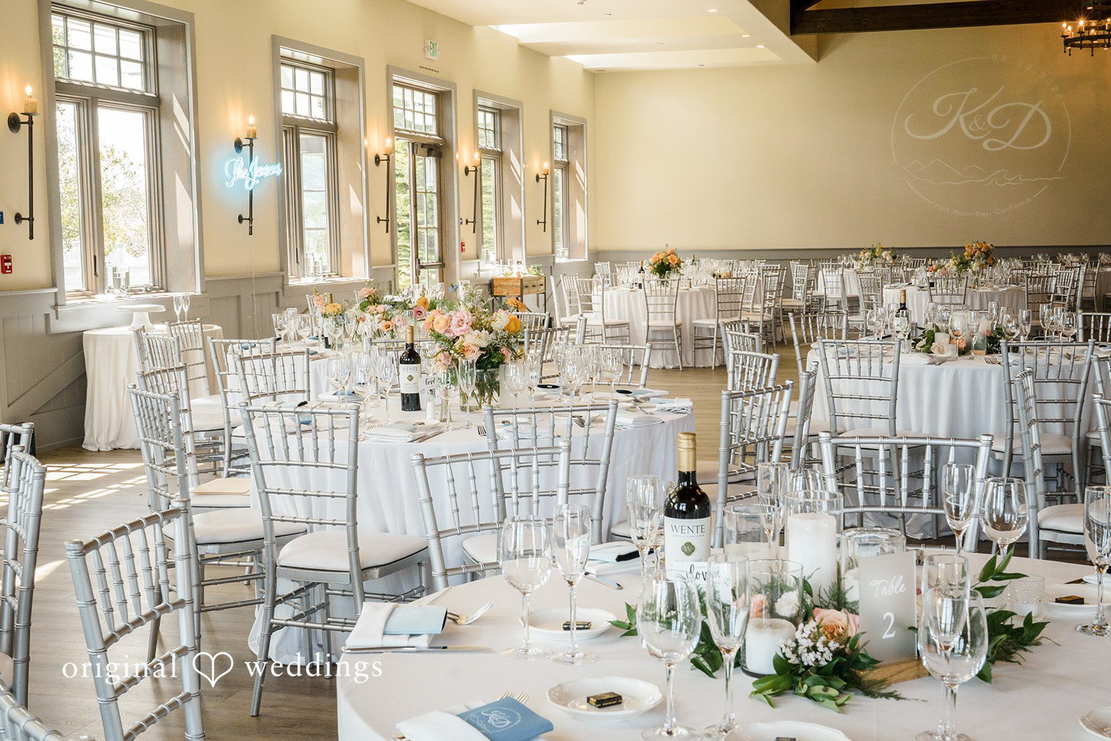 Daniel + Karen An empty reception hall filled with round tables, white chairs, and elegant table settings.