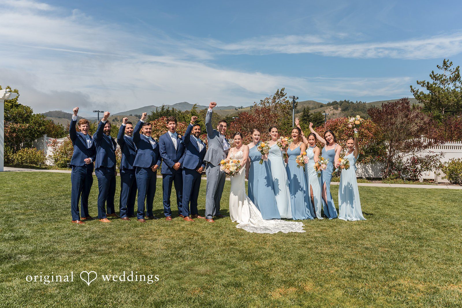 Daniel + Karen A large wedding party posing together on a grassy lawn with a scenic backdrop.