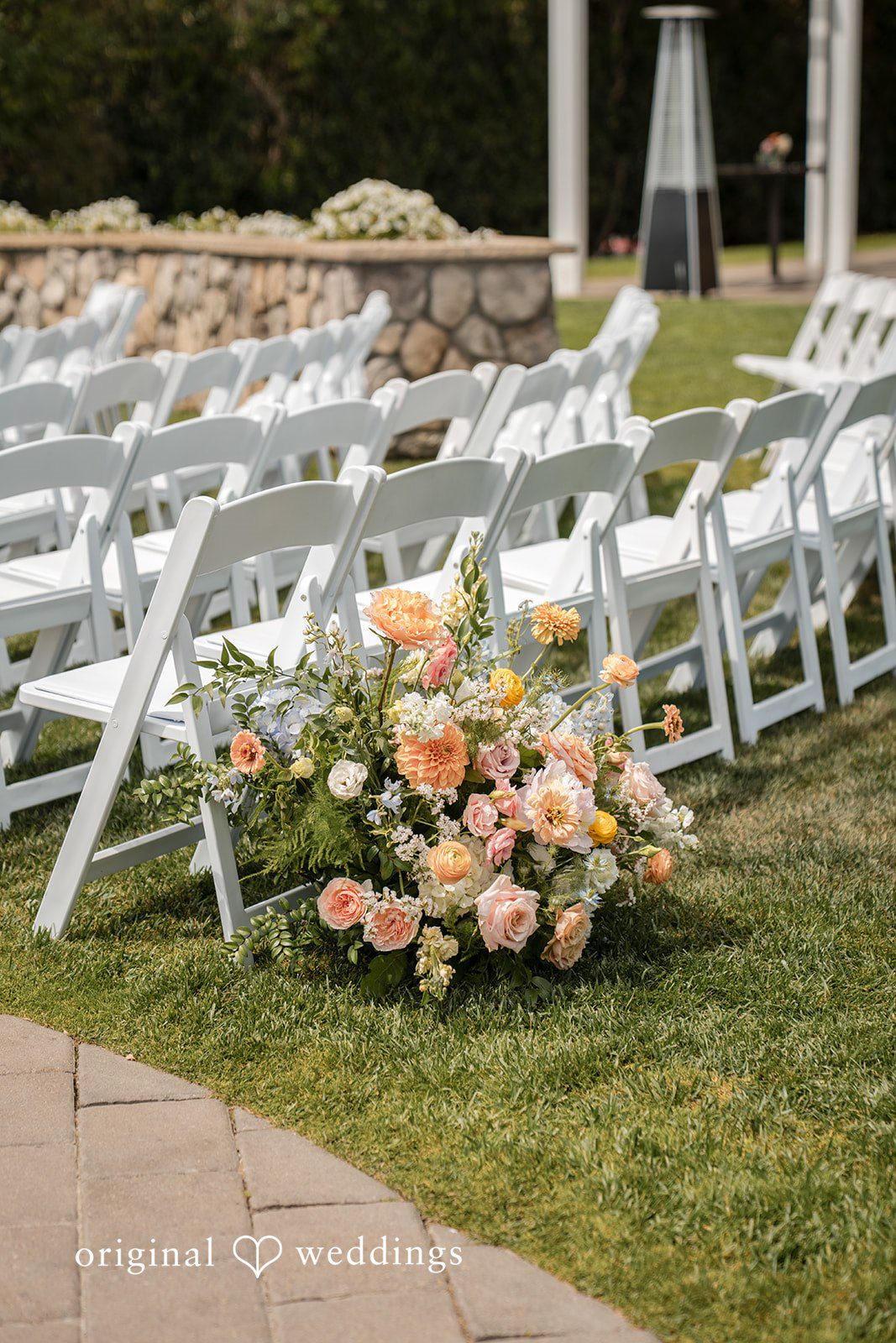 Daniel + Karen White folding chairs arranged on a lawn with a peach and pink floral display in the foreground.