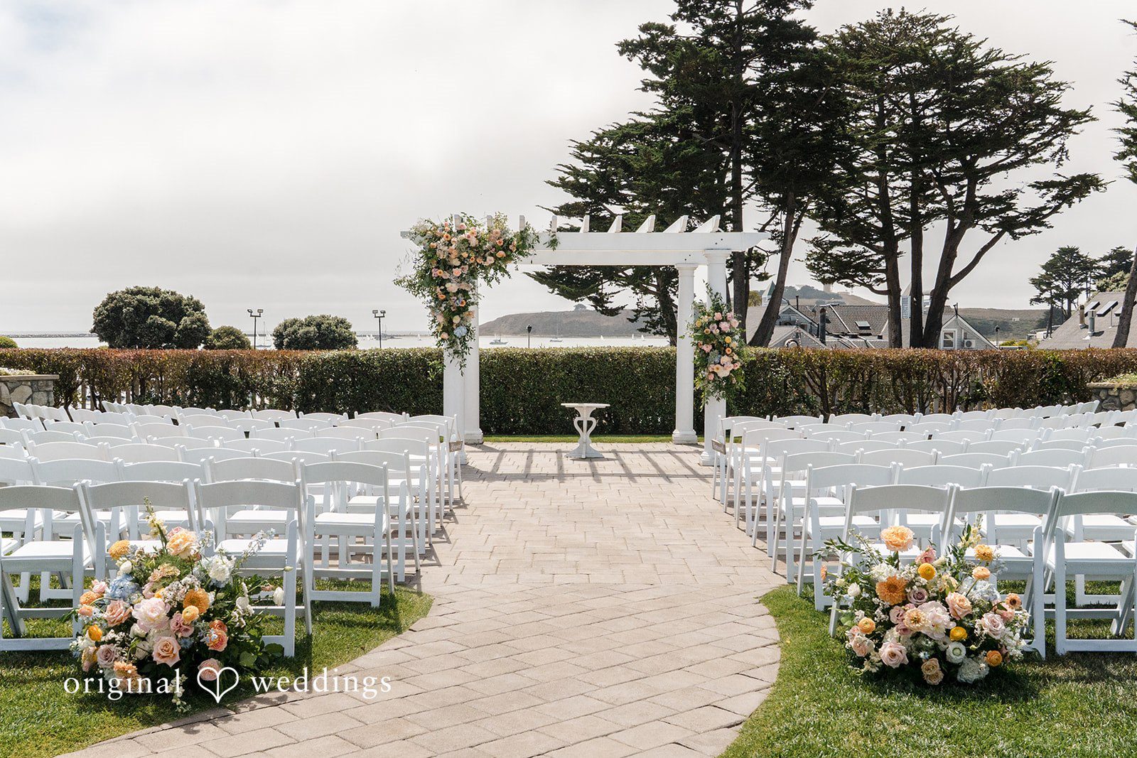 Daniel + Karen An empty wedding ceremony aisle lined with floral arrangements leading to a wooden arbor.