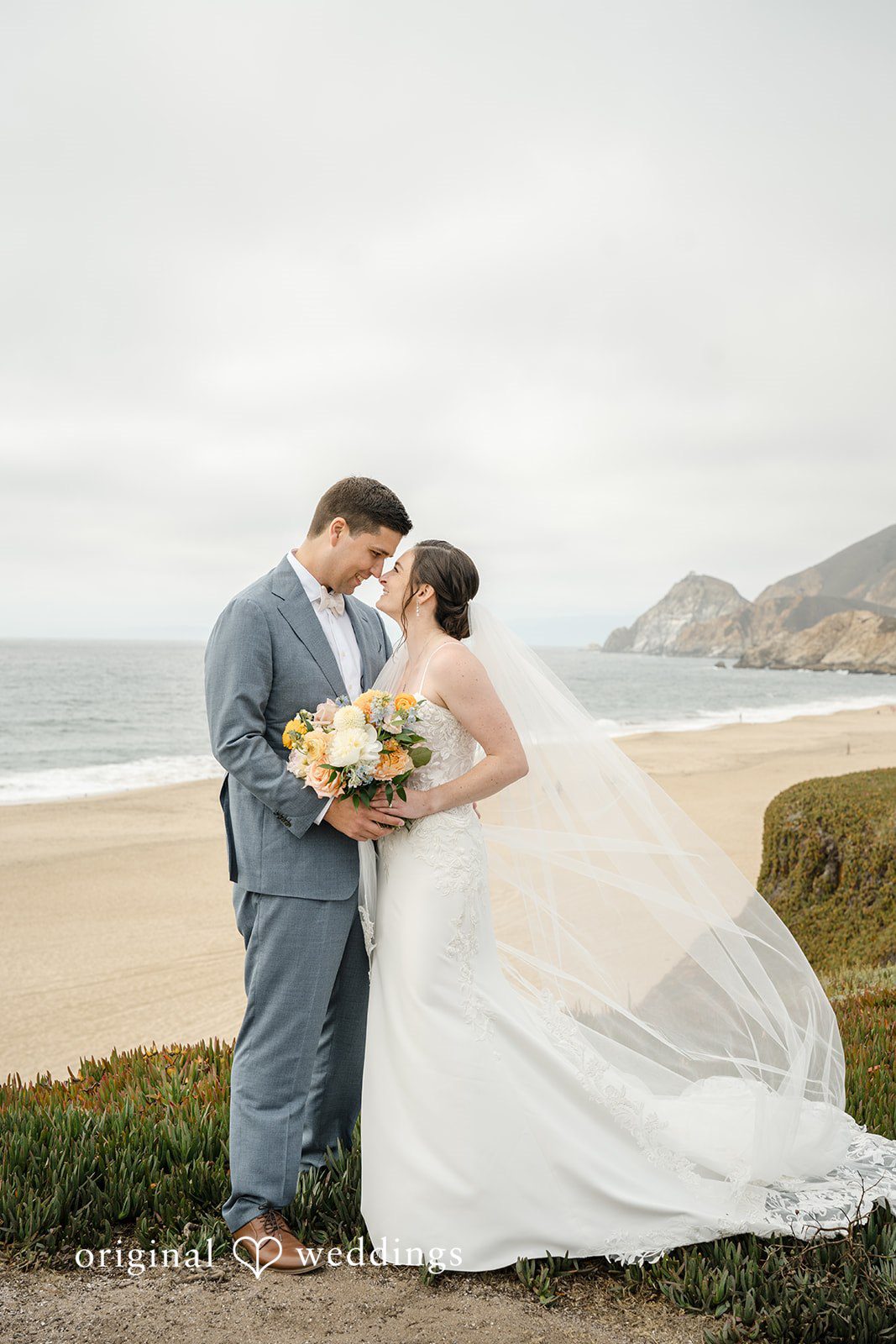 Daniel + Karen A groom looking at his bride as her long wedding dress train flows behind them on the beach.