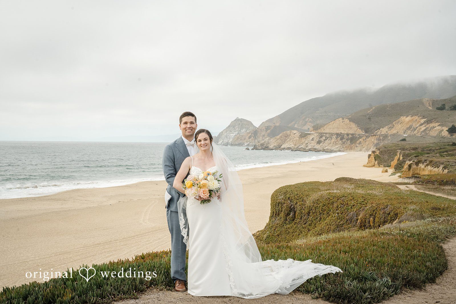 Daniel + Karen A bride standing alone on a beach pathway with her long veil trailing behind.