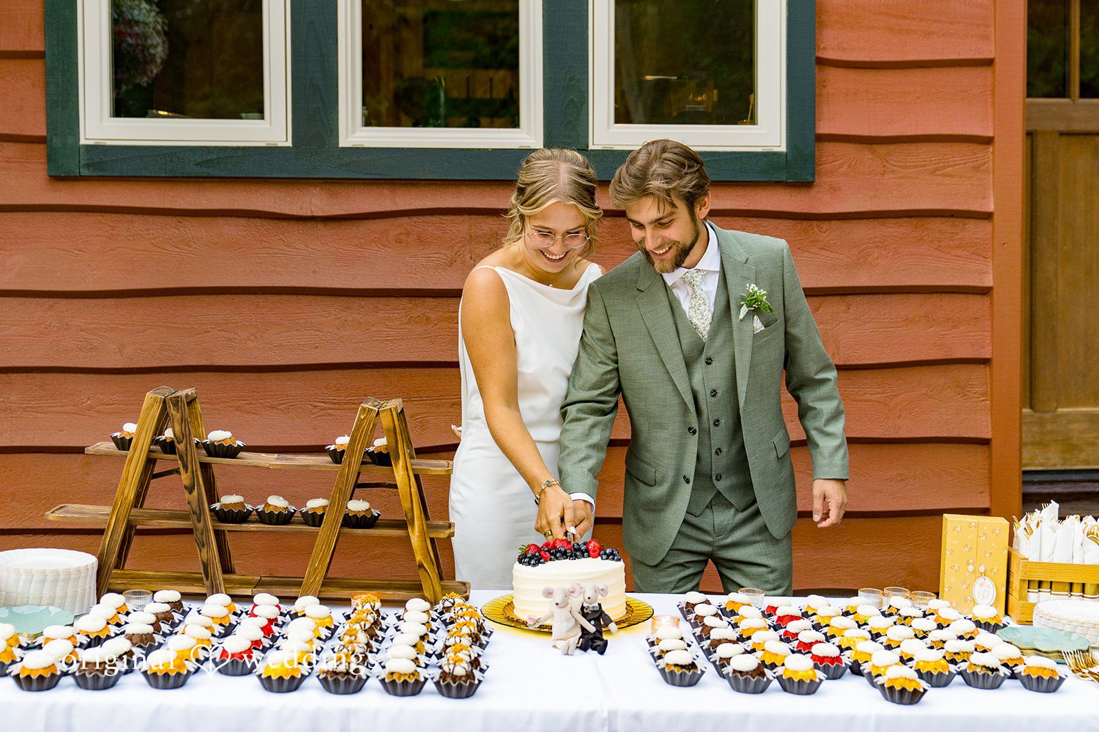 Abigail + Reece A bride and groom cutting wedding cake and looking at a dessert table filled with cupcakes.