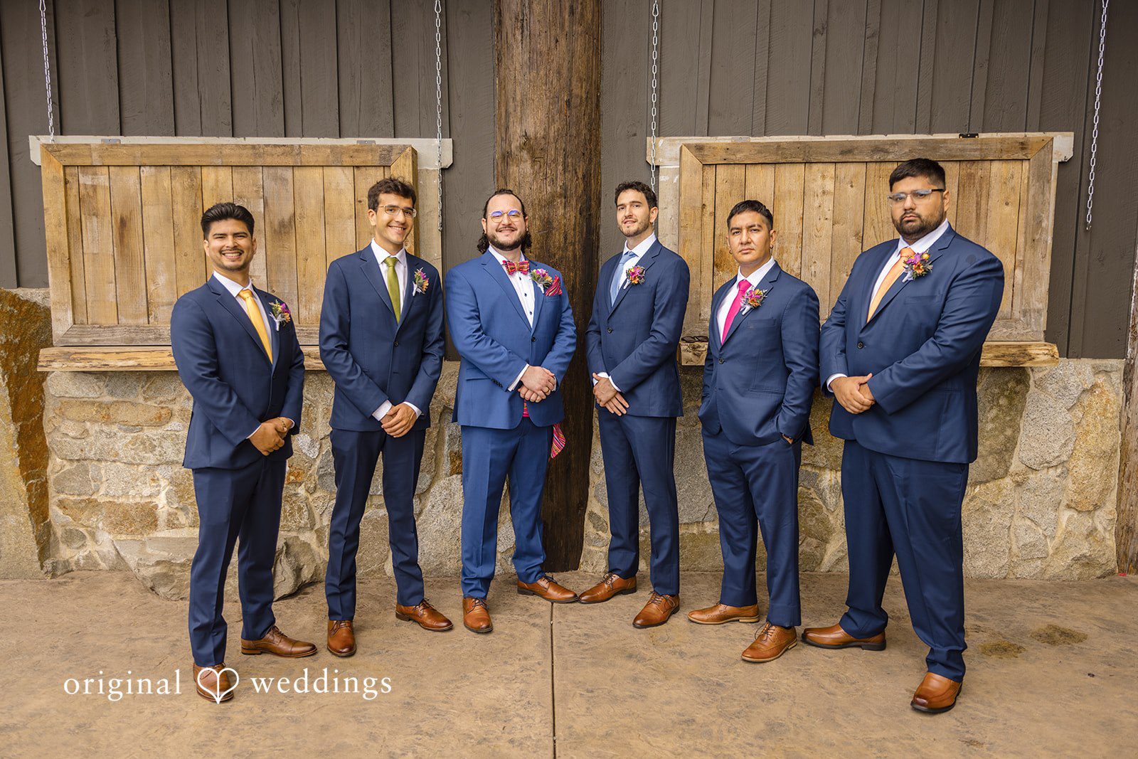 Diego + Adel A group of groomsmen in matching blue suits posing together in front of a rustic wooden structure.