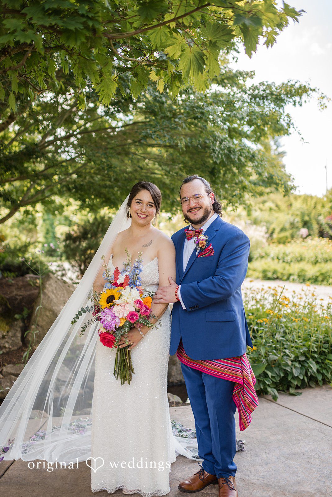 Diego + Adel A bride and groom walking together along a garden pathway with guests in the background.