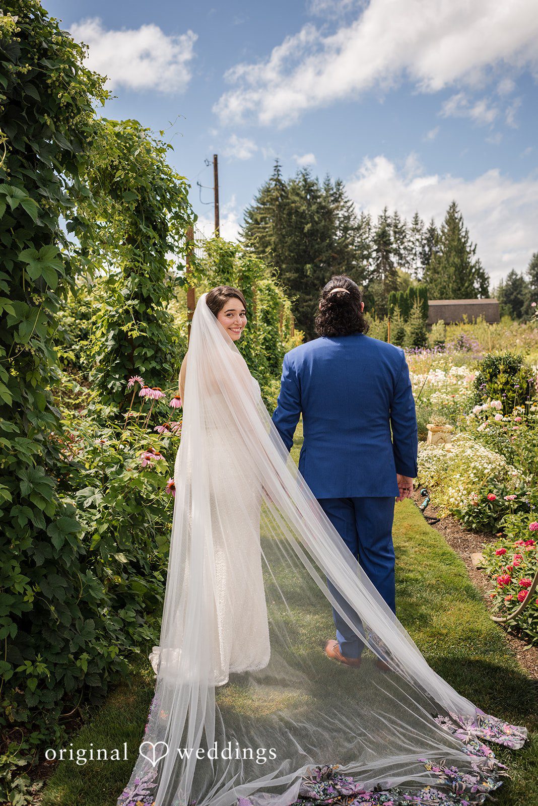 Diego + Adel A bride with a long trailing veil standing next to her groom in an open field.