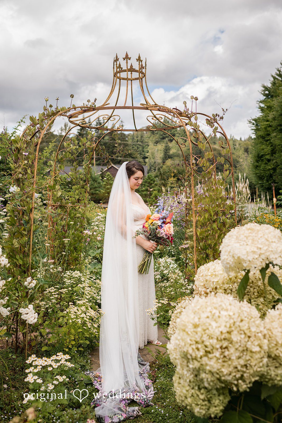 Diego + Adel A bride walking through a lush garden path lined with large white flower bushes.