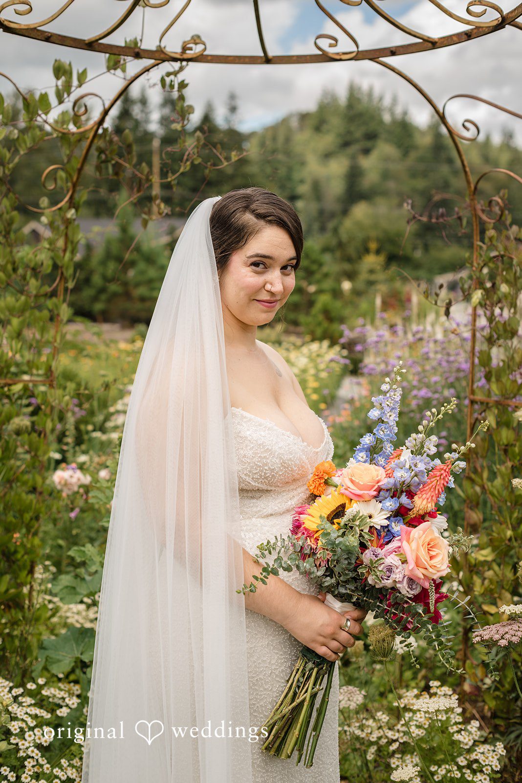 Diego + Adel A bride with a long sheer veil holding a bright bouquet in a garden setting.