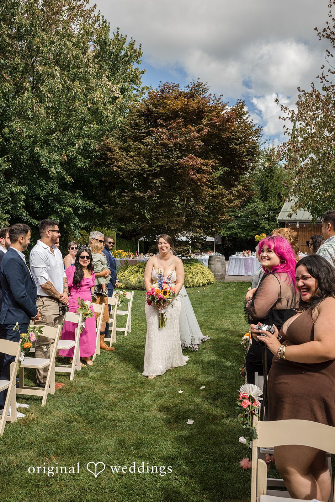 Diego + Adel Bride walking down an aisle and wedding guests seated on chairs under trees watching an event.