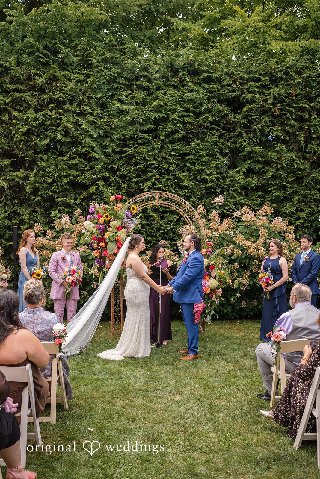 Diego + Adel A wide shot of a wedding ceremony in progress before a large green hedge and floral arch.
