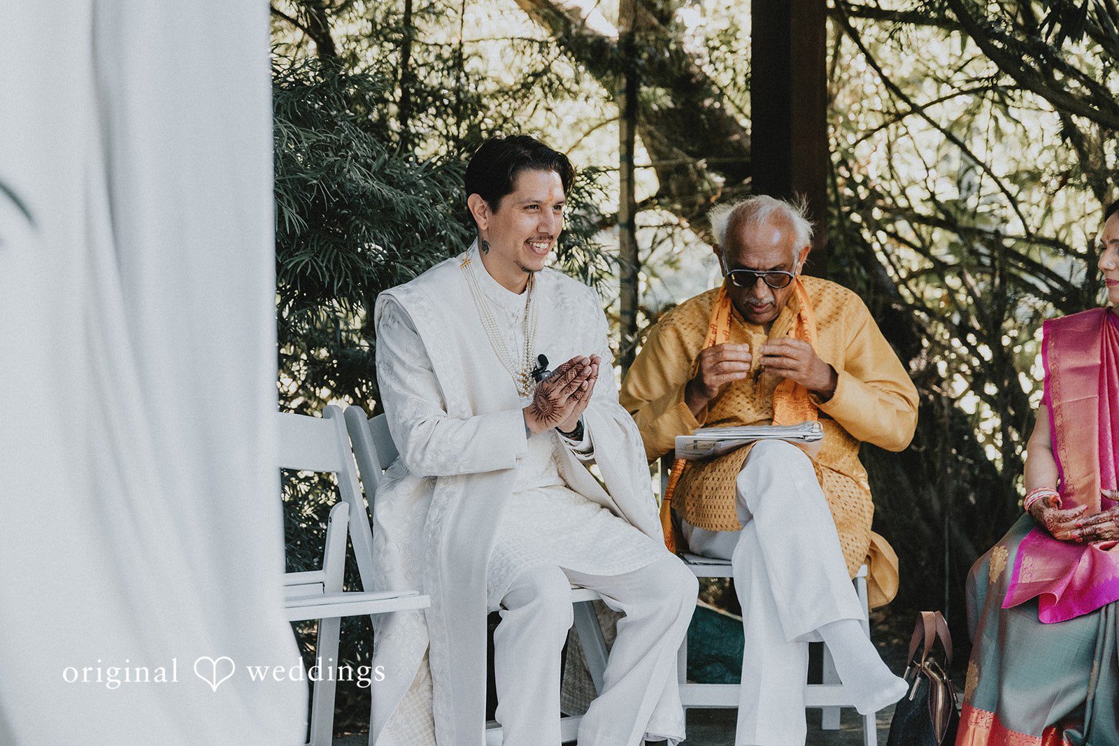 A smiling man in a white sherwani sits with his hands pressed together next to an older man wearing a yellow kurta.